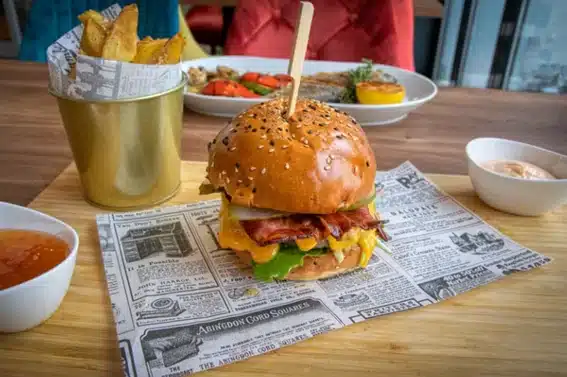 Hamburguesa gourmet con bacon y queso sobre papel antigrasa estilo periódico junto a patatas y salsas