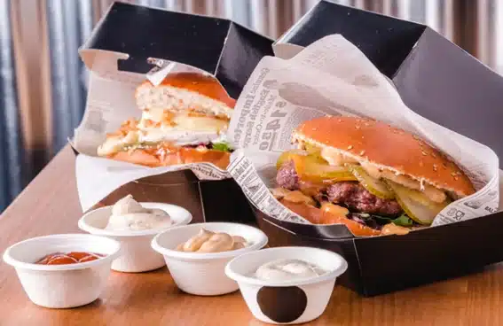 Dos hamburguesas en cajas de cartón negras con papel antigrasa y salseros de ketchup y mayonesa